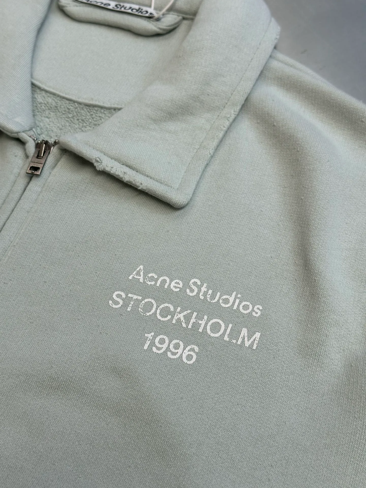Ac vintage collared zipper sweatshirt uses 32-count top layer and 40-count bottom layer of 3-strand composite fleece structure, with a weight of g/㎡. The 32-count yarn on the top layer is fine and smooth, giving the surface a smooth touch and excellent anti-pilling properties; the 40-count 3-ply yarn on the bottom layer is treated with a raising process to form a rich and soft fluff layer, providing excellent warmth and skin-friendly wrapping. With 21 2×2 ribs, the g/㎡ weight ensures that the collar, cuffs and hem have excellent tensile fatigue resistance and shape retention capabilities. During piece dyeing, the entire fabric is immersed in the dye vat after weaving, and the dye molecules fully penetrate into the fibers, ensuring full, deep colors and excellent color fastness. Each batch of dyeing produces subtle differences, making the color tone of the single product unique. On this basis, the paint is sprayed irregularly on the surface of the garment through manual spray painting to create a splash-like retro effect. The distressed processing of holes causes the color to fade and mottle in parts. The ribbed trim is also treated to form slight whitening and fluffing, which visually echoes the spray-painted effect on the body, making the new clothes appear as if they have been worn for a long time and exposed to the sun. The front logo stamp uses a slurry printing process. The printing slurry contains a special microcapsule foaming agent. After being baked at high temperature, the slurry layer expands to form three-dimensional bulges, and the surface presents a granular texture similar to inkjet paint. This process simulates the effect of random drops of pigment after being atomized during spray gun spraying. The edges of the pattern present a natural feathering transition, rather than the rigid boundaries of glue printing. The printing surface has a fine graininess and soft elasticity to the touch, forming a closed loop of the process with the distressed effect of the spray painting on the body. size：xs s m l